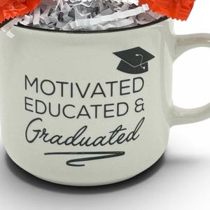 May include: Cream-colored ceramic mug with the words "MOTIVATED EDUCATED & Graduated" in black script and a graduation cap graphic. The mug is filled with shredded paper and wrapped candies.