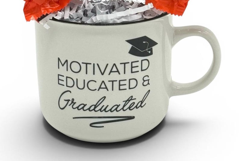 May include: Cream-colored ceramic mug with the words "MOTIVATED EDUCATED & Graduated" in black script and a graduation cap graphic. The mug is filled with shredded paper and wrapped candies.