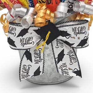 May include: A graduation gift basket filled with candy and decorated with a white ribbon printed with black graduation caps and the words "Congrats GRADI". The basket is in a galvanized metal bucket.