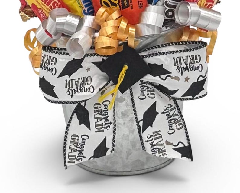 May include: A graduation gift basket filled with candy and decorated with a white ribbon printed with black graduation caps and the words "Congrats GRADI". The basket is in a galvanized metal bucket.