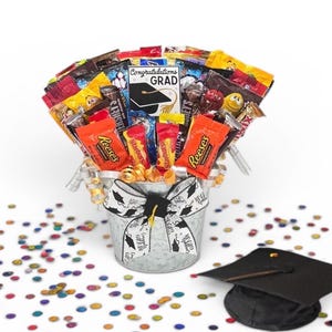 May include: A graduation-themed candy bouquet in a galvanized metal bucket, filled with various wrapped candies. The bucket is adorned with a graduation ribbon. A graduation cap and colorful confetti are scattered around the bucket.