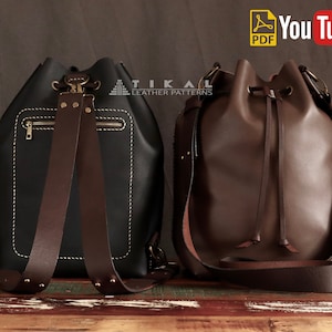 May include: Two leather bucket bags with brown straps. The bag on the left is black with a zipped pocket and a brown strap. The bag on the right is brown with a drawstring closure and a brown strap. Both bags are on a wooden surface.