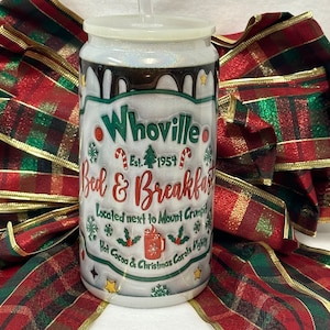May include: A clear, glittery tumbler with a lid and straw, featuring the text "Whoville Bed & Breakfast" with Christmas-themed graphics. The design includes snowflakes, candy canes, and a mug of hot cocoa. The tumbler is set against a backdrop of a red and green plaid ribbon.