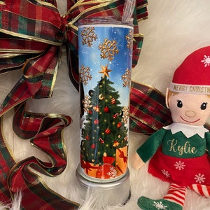 May include: A festive Christmas tumbler with a seasonal design. The tumbler displays a decorated Christmas tree, wrapped gifts, and gold snowflakes against a blue sky. A red and green plaid ribbon and an elf doll are also in the image.
