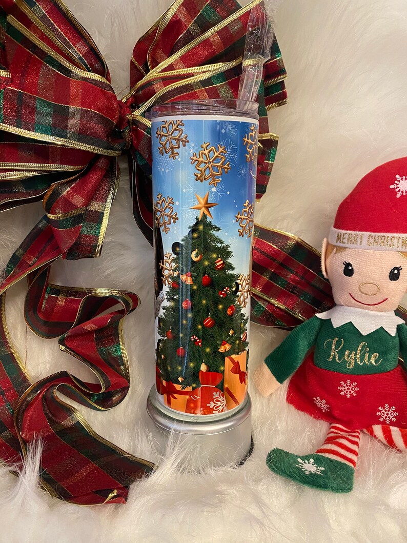 May include: A festive Christmas tumbler with a seasonal design. The tumbler displays a decorated Christmas tree, wrapped gifts, and gold snowflakes against a blue sky. A red and green plaid ribbon and an elf doll are also in the image.