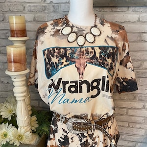May include: A mannequin displays a cream-colored t-shirt with a brown and blue cowhide pattern and the words "Wranglin Mama". The shirt is paired with a matching skirt and a studded belt with a large, sparkling buckle. A white stone necklace completes the look.