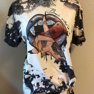 May include: White t-shirt with a black and white tie-dye pattern. The shirt features a graphic of blue lips, a hand with tattoos, and the words "Basic witch". A silver necklace is around the neck.