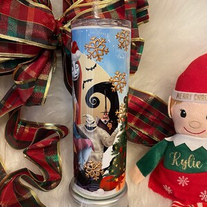 May include: A stainless steel tumbler featuring characters from The Nightmare Before Christmas. The design includes Sally, Jack Skellington, and Oogie Boogie, with snowflakes and a Christmas tree. The tumbler is decorated with a red and green plaid ribbon.