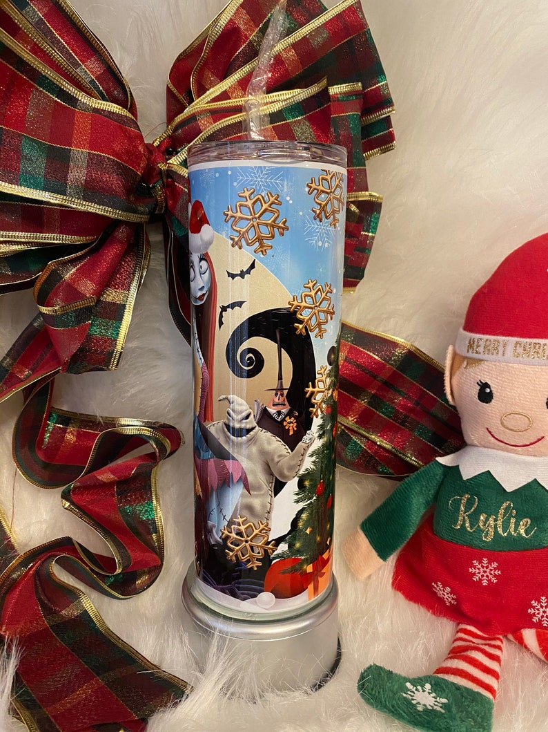 May include: A stainless steel tumbler featuring characters from The Nightmare Before Christmas. The design includes Sally, Jack Skellington, and Oogie Boogie, with snowflakes and a Christmas tree. The tumbler is decorated with a red and green plaid ribbon.