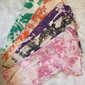 May include: Five long-sleeved t-shirts in various colors, including green, orange, purple, black and white, and pink. Each shirt has a tie-dye or bleach-dye pattern. The shirts are arranged on a white, fluffy surface.