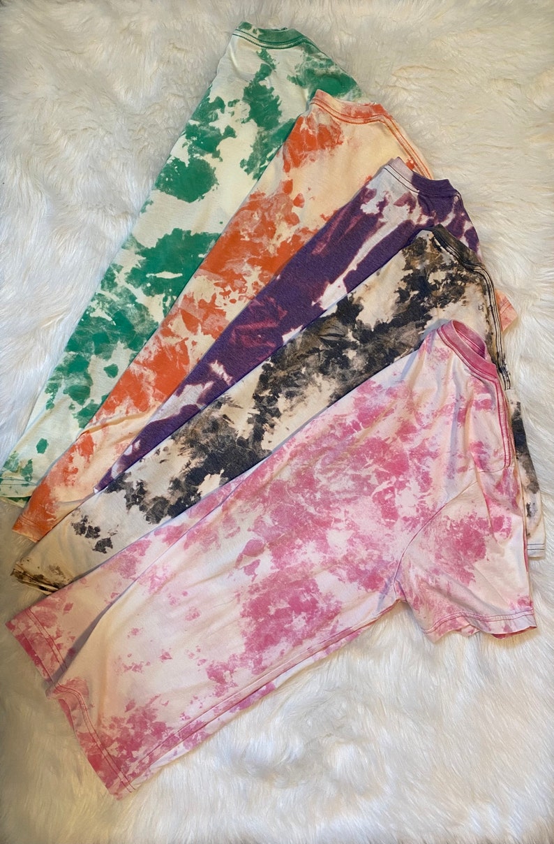 May include: Five long-sleeved t-shirts in various colors, including green, orange, purple, black and white, and pink. Each shirt has a tie-dye or bleach-dye pattern. The shirts are arranged on a white, fluffy surface.