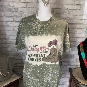 May include: Olive green t-shirt with a bleached design. The shirt features the text "My Daughter wears Combat Boots" with an illustration of combat boots and flowers. A necklace and jewelry are also visible.