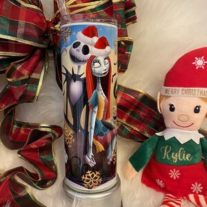 May include: A stainless steel tumbler featuring Jack Skellington and Sally from The Nightmare Before Christmas, wearing Santa hats. The tumbler has a clear straw and is decorated with snowflakes. A red and green plaid ribbon and an elf doll are in the background.