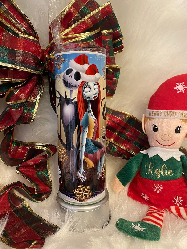 May include: A stainless steel tumbler featuring Jack Skellington and Sally from The Nightmare Before Christmas, wearing Santa hats. The tumbler has a clear straw and is decorated with snowflakes. A red and green plaid ribbon and an elf doll are in the background.
