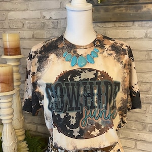 May include: A bleached t-shirt with the words "COWHIDE junkie" in a distressed design. The shirt is paired with a turquoise stone necklace and a studded belt. The shirt has a brown, black, and white pattern.