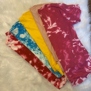 May include: A collection of five t-shirts in various colors, including red, blue, yellow, tan, and maroon. The red and blue shirts have a white, tie-dye pattern. The shirts are arranged on a white, fluffy surface.