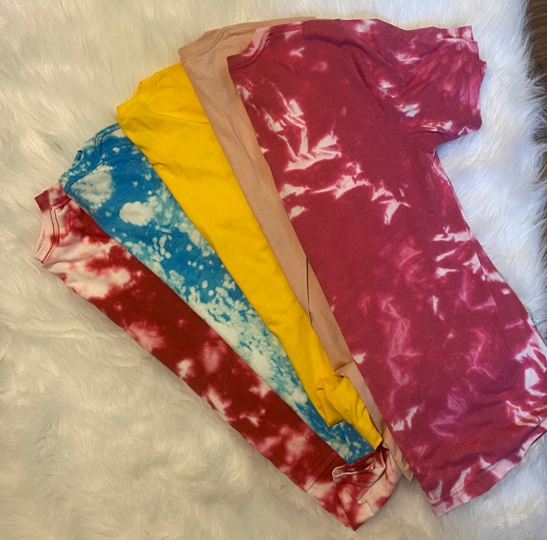 May include: A collection of five t-shirts in various colors, including red, blue, yellow, tan, and maroon. The red and blue shirts have a white, tie-dye pattern. The shirts are arranged on a white, fluffy surface.