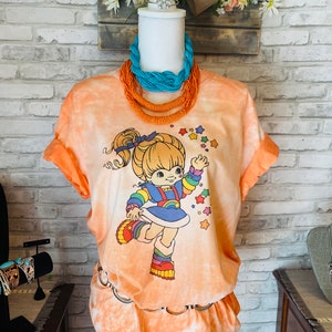 May include: An orange tie-dye t-shirt and skirt set with a cartoon character graphic. The character is wearing a rainbow outfit and surrounded by stars. A turquoise and orange beaded necklace is displayed on a mannequin.