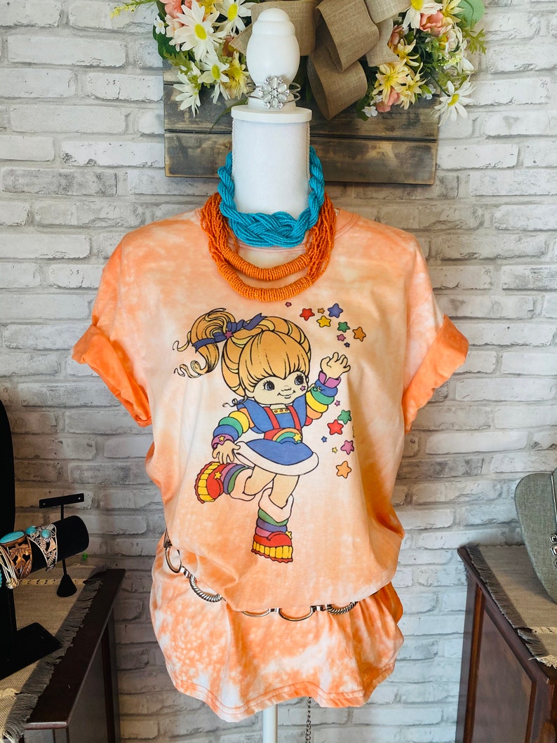 May include: An orange tie-dye t-shirt and skirt set with a cartoon character graphic. The character is wearing a rainbow outfit and surrounded by stars. A turquoise and orange beaded necklace is displayed on a mannequin.