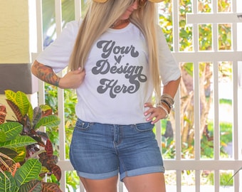 May include: A white t-shirt with the text "Your Design Here" in a retro font. The shirt is being worn by a person with long blonde hair and a straw hat. The person is standing in front of a white picket fence.