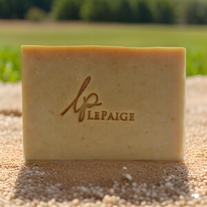 Puede incluir: Una pastilla de jabón rectangular de color marrón claro, sobre una superficie arenosa. El jabón tiene el texto "lp LEPAIGE" en relieve, en un color marrón más oscuro. El fondo muestra un campo verde borroso y árboles.