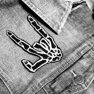 May include: A black and white skeleton hand patch in the shape of a rock and roll hand gesture. The patch is sewn onto a denim jacket.