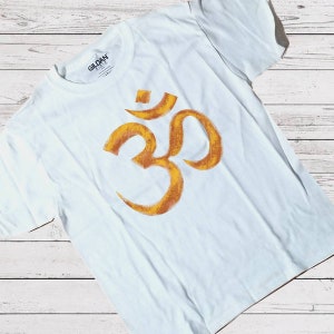 May include: A white t-shirt with a gold Om symbol printed on the front. The symbol is a stylised Sanskrit script.