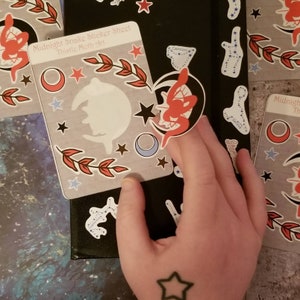 May include: A sticker sheet featuring a range of celestial designs, including moons, stars, and a snake. The sheet is labelled "Midnight Snake Sticker Sheet Thistle Moth Art".