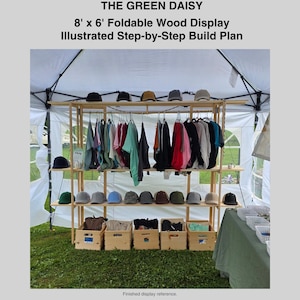 Puede incluir: Un expositor de madera clara con tres estantes horizontales y una barra superior. La estructura sostiene gorras de béisbol en los estantes, prendas colgadas de la barra y cajas de madera en la base. Texto: THE GREEN DAISY 8' x 6' Foldable Wood Display Illustrated Step-by-Step Build Plan.