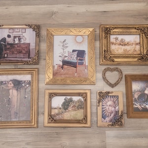 May include: A collection of framed art pieces with ornate gold frames. The artwork includes a variety of images, such as a woman, a landscape, and a still life. The frames are arranged on a light wood-grain surface.
