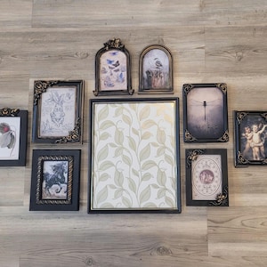 May include: A collection of framed art pieces in various sizes and styles. The frames are black with gold accents, and the artwork includes botanical prints, butterflies, and angel figures. The arrangement is displayed on a light wood-grain surface.