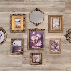 May include: A collection of ornate, gold-framed pictures and mirrors arranged on a light wood-grain wall. The frames vary in shape and size, with some containing landscape paintings, floral arrangements, and portraits. The mirrors have decorative frames.