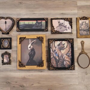 May include: An assortment of framed art pieces and a hand mirror are arranged on a wooden surface. The art includes a heart-shaped frame, a black cat, a rabbit figure, and a cat mosaic. The frames have ornate gold and black detailing.