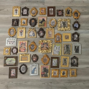 May include: A collection of ornate, vintage-style picture frames in various shapes and sizes. The frames are gold, silver, and brown, with images of cats, animals, and abstract designs. The frames are arranged on a light gray wooden surface.