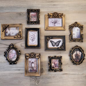 May include: A collection of nine ornate picture frames with various designs and colors, including gold, black, and white. The frames are arranged in a grid pattern on a light wood surface. Some frames contain images of flowers, butterflies, and other nature-inspired designs.
