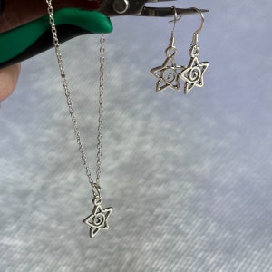 Op de afbeelding: Een zilveren ketting- en oorstekerset met een sterhanger met een swirl-ontwerp. De ketting heeft een delicate ketting en de oorbellen hebben een eenvoudige haaksluiting.