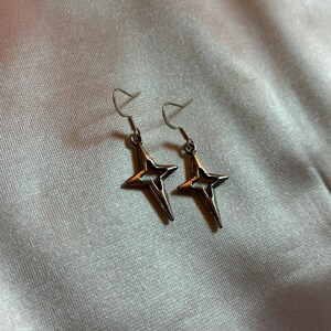 May include: A pair of silver-colored star-shaped earrings. Each earring features a four-pointed star design with a cutout center and a hook for pierced ears. The earrings are displayed against a light-colored, textured background.