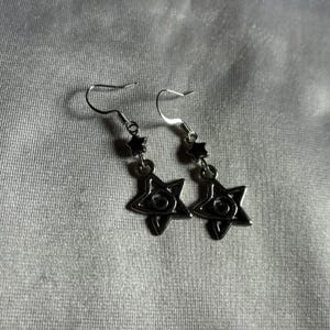 May include: A pair of silver earrings with a star-shaped charm featuring a black eye design. The earrings have a small black star charm above the eye charm.