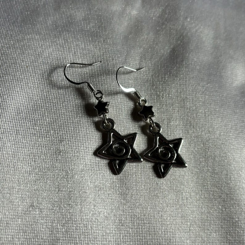 May include: A pair of silver earrings with a star-shaped charm featuring a black eye design. The earrings have a small black star charm above the eye charm.