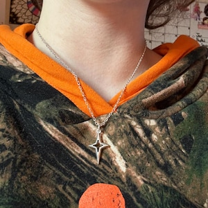 May include: A silver-colored necklace featuring a four-pointed star pendant. The pendant has a hollow center and hangs from a delicate chain. The necklace is displayed against a camouflage-patterned hoodie with an orange hood lining.