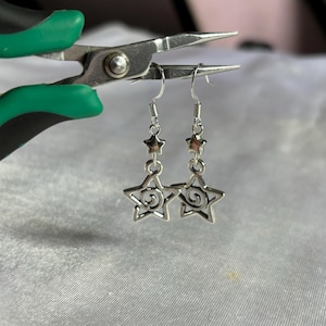 May include: Silver dangle earrings with a star design. The star has a swirl pattern inside. The earrings are hanging from a silver hook.