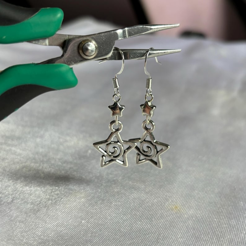 May include: Silver dangle earrings with a star design. The star has a swirl pattern inside. The earrings are hanging from a silver hook.