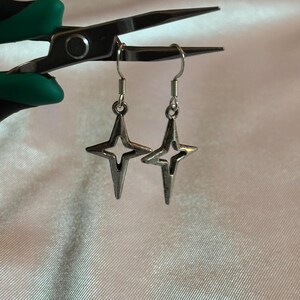 May include: A pair of silver-colored star-shaped earrings with a four-pointed design. The earrings are suspended from silver hooks. The earrings are displayed against a white, textured background, held by a pair of pliers with green handles.