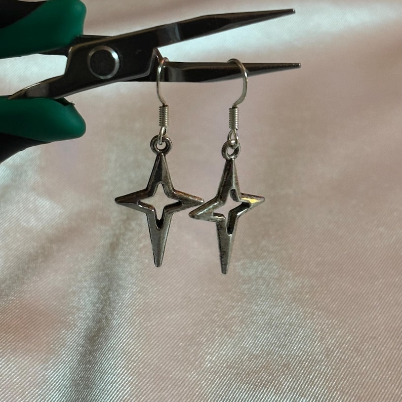 May include: A pair of silver-colored star-shaped earrings with a four-pointed design. The earrings are suspended from silver hooks. The earrings are displayed against a white, textured background, held by a pair of pliers with green handles.