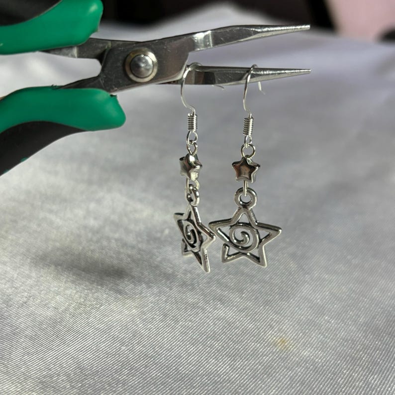 May include: A pair of silver star-shaped earrings with a swirl design. The earrings are hanging from a silver chain with a small star charm at the top.
