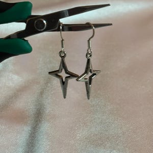 May include: A pair of silver-colored star-shaped earrings with a four-pointed design. The earrings are suspended from silver hooks. The background is a soft, off-white fabric. The earrings are held by a pair of scissors.