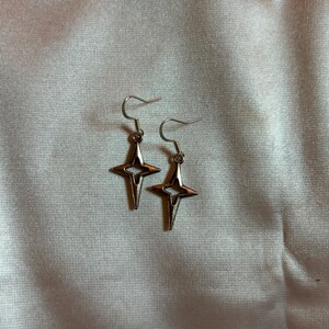 May include: A pair of silver-colored star-shaped earrings with a hook closure. The earrings have a four-pointed star design with a cutout in the center. The earrings are displayed on a light-colored, textured surface.