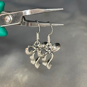 May include: A pair of silver bow earrings with a simple design. The earrings are hanging from a pair of pliers.