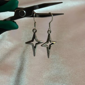 May include: A pair of silver-toned star-shaped earrings with a four-pointed design. The earrings are suspended from silver hooks. The background is a soft, off-white fabric, and the earrings are held by a pair of pliers with green handles.