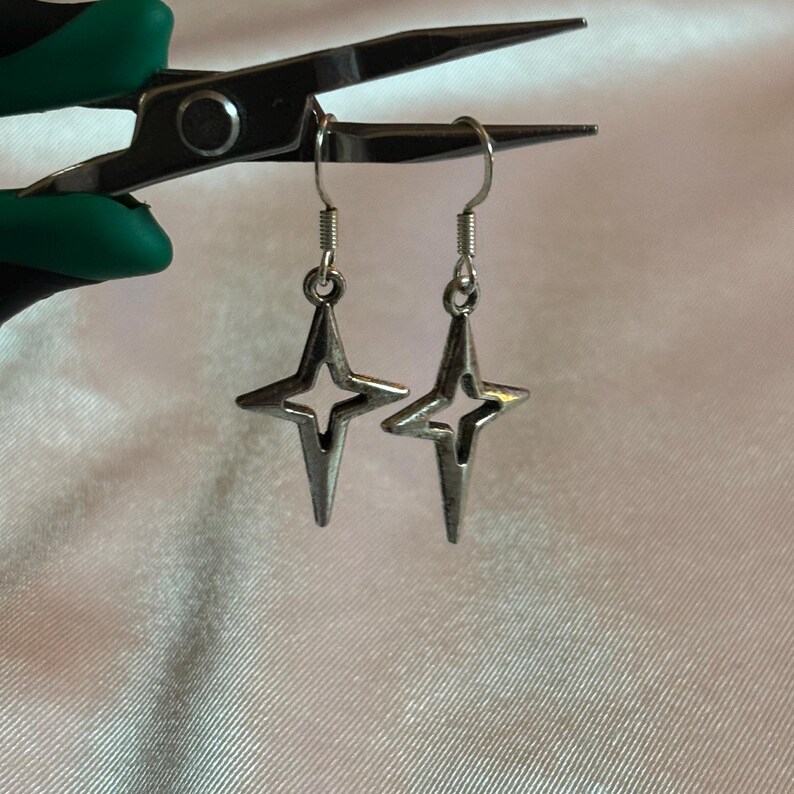 May include: A pair of silver-toned star-shaped earrings with a four-pointed design. The earrings are suspended from silver hooks. The background is a soft, off-white fabric, and the earrings are held by a pair of pliers with green handles.
